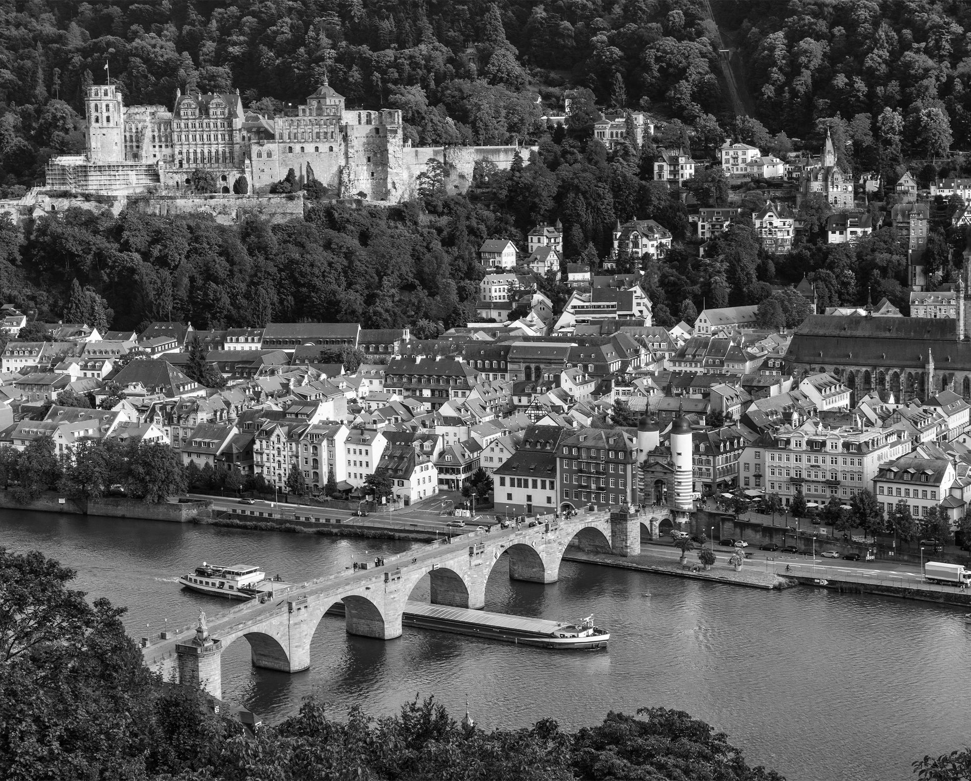 Monochrome Ansicht der Stadt Heidelberg mit der Alten Brücke über den Neckar, historischen Gebäuden, der Heiliggeistkirche und dem Heidelberger Schloss vor einer bewaldeten Hügellandschaft.