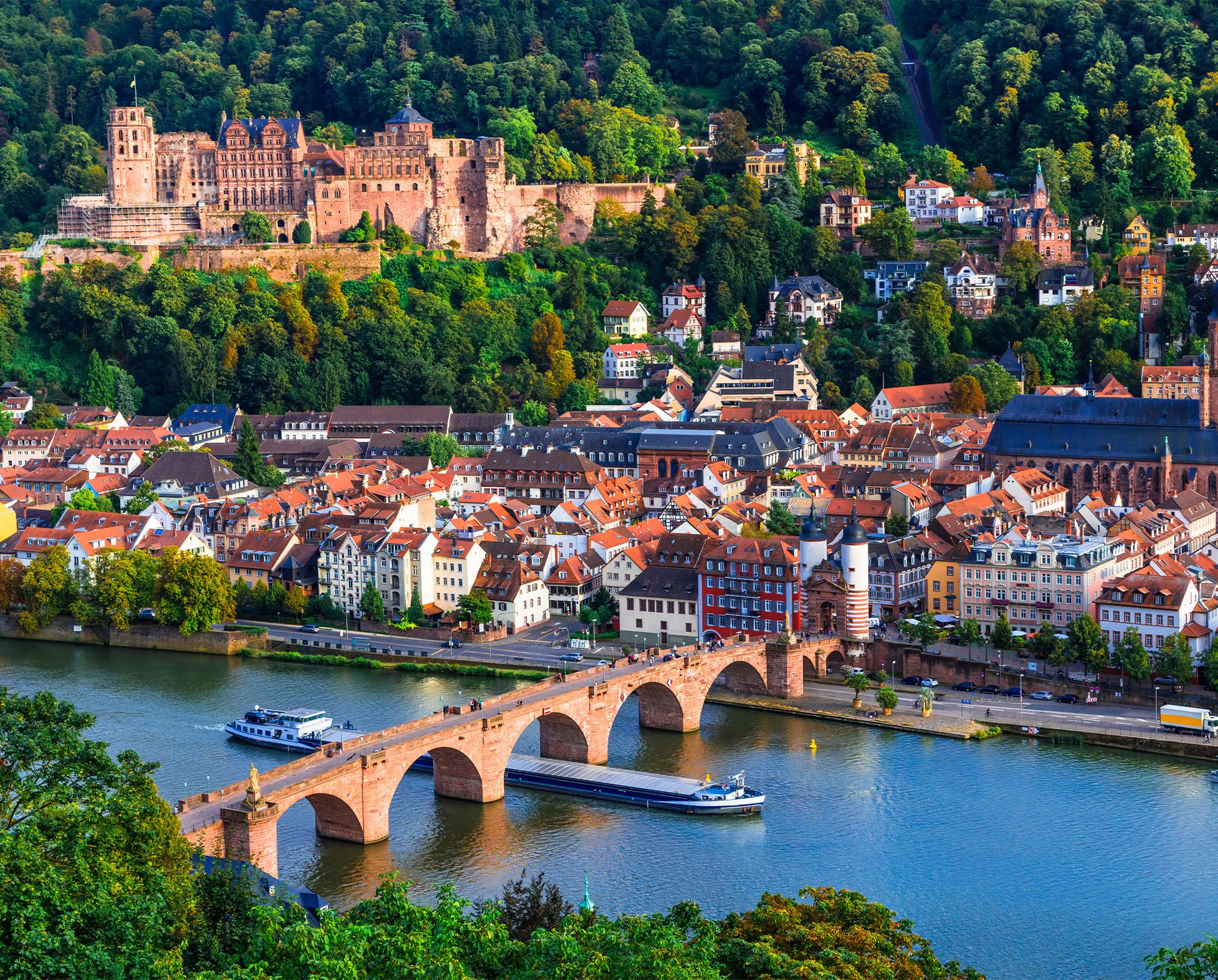 View of the city of Heidelberg with the Old Bridge over the Neckar, historical buildings, the Church of the Holy Spirit and the Heidelberg Castle in front of a wooded hilly landscape.